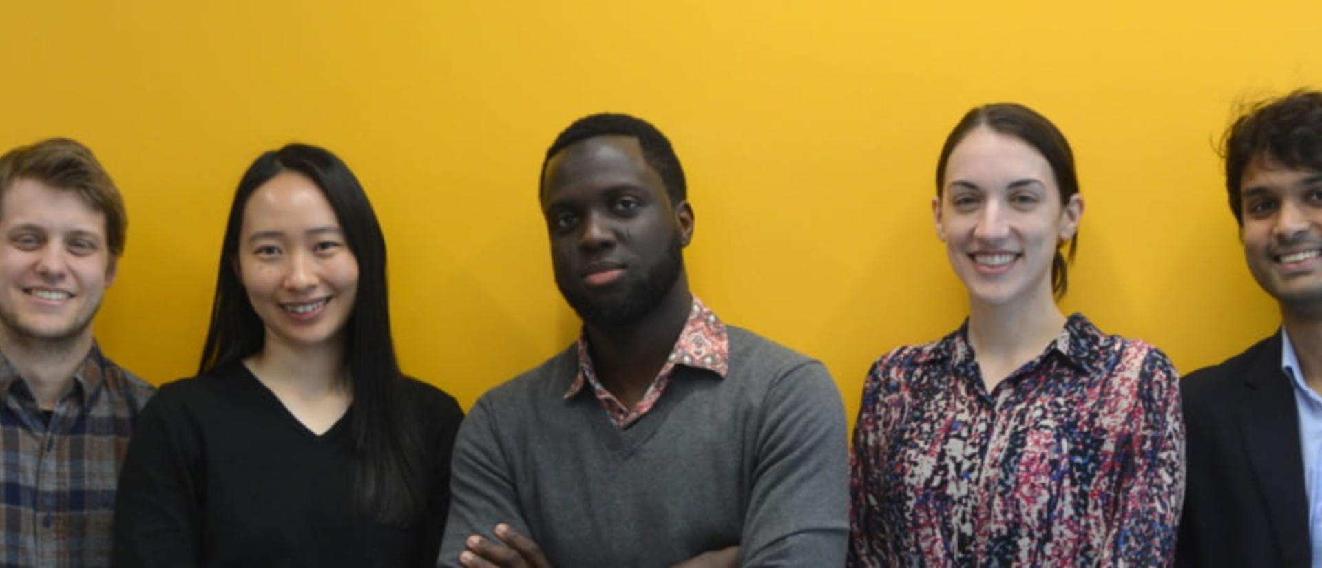Photo of Rowland Institute's Current Fellows. A diverse group of seven people stands smiling in front of a yellow background. Their expressions convey friendliness and confidence, suggesting a positive team dynamic.