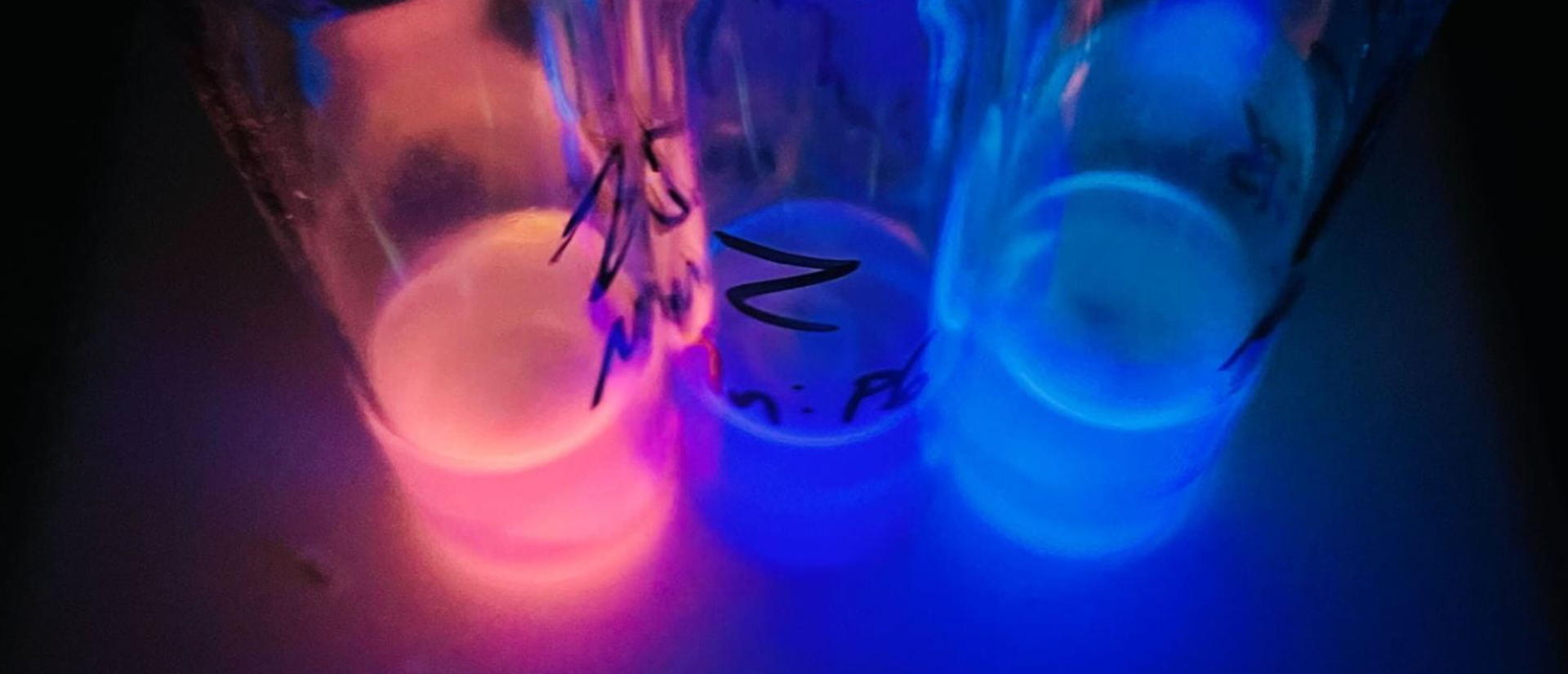 Five glass vials glow in dark hues of pink and blue under ultraviolet light, creating a mysterious and vibrant atmosphere.